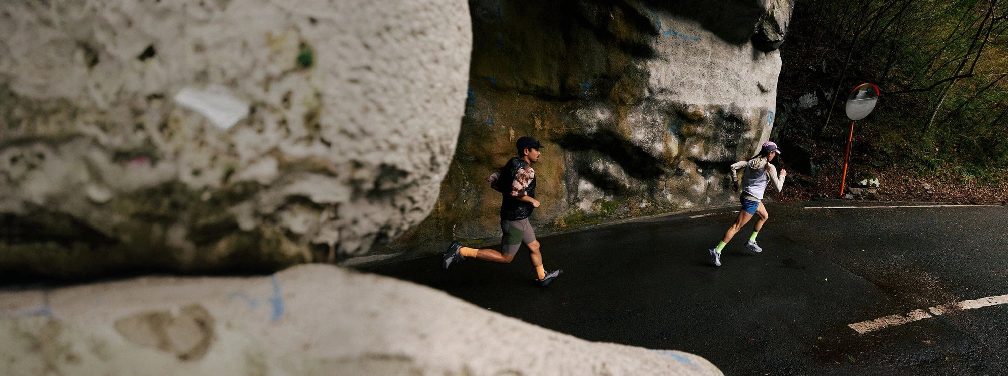 A man and woman running pair emerging mid-stride from a rock tunnel on a paved forest road.