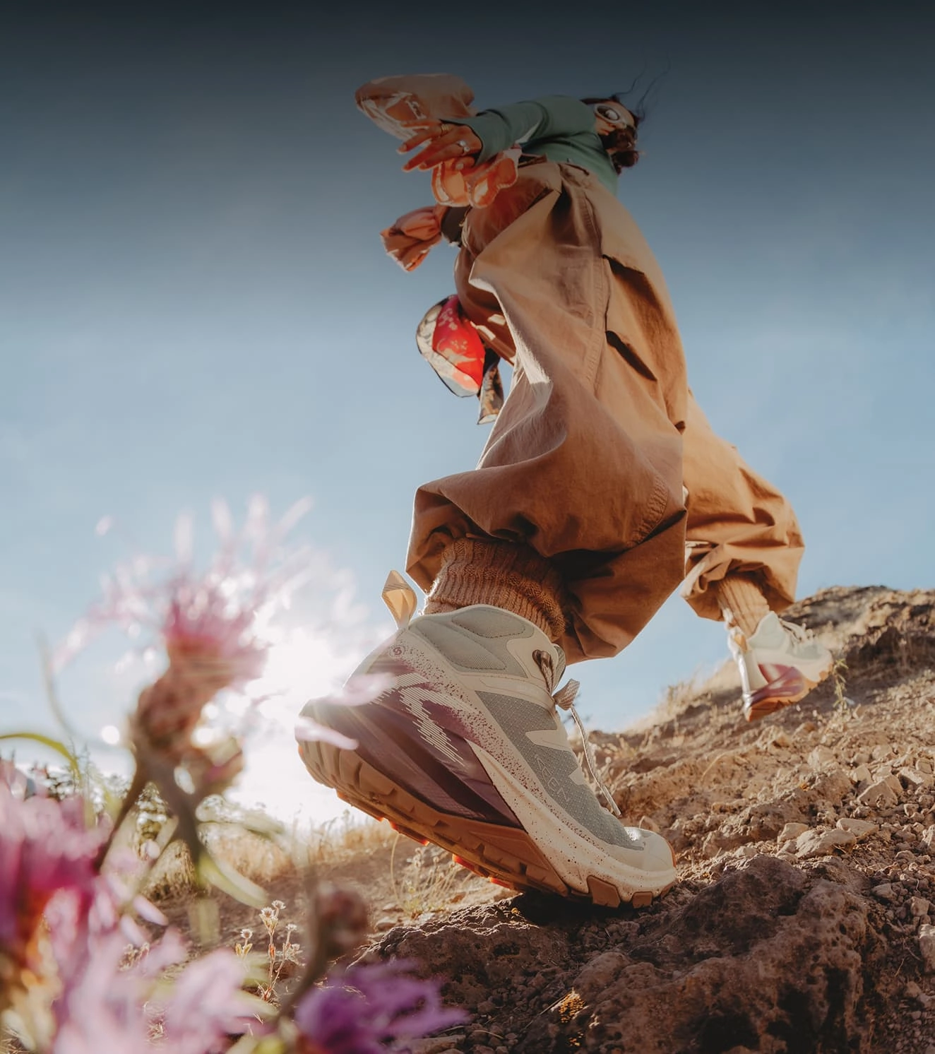 A woman in orange baggy hiking pants walking up a steep dirt path lined by purple flowers while wearing Targhee Apex Hiking boots.