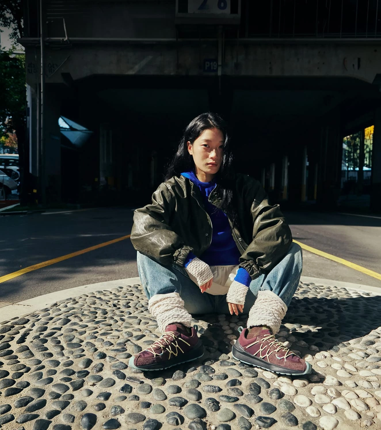 A person in stylish streetwear sitting in the middle of a city street while wearing maroon and green Uneek Trekker sneakers.