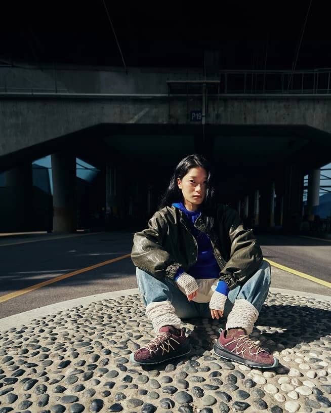 A person in stylish streetwear, sitting in the middle of a city street while wearing maroon and green Uneek Trekker sneakers.