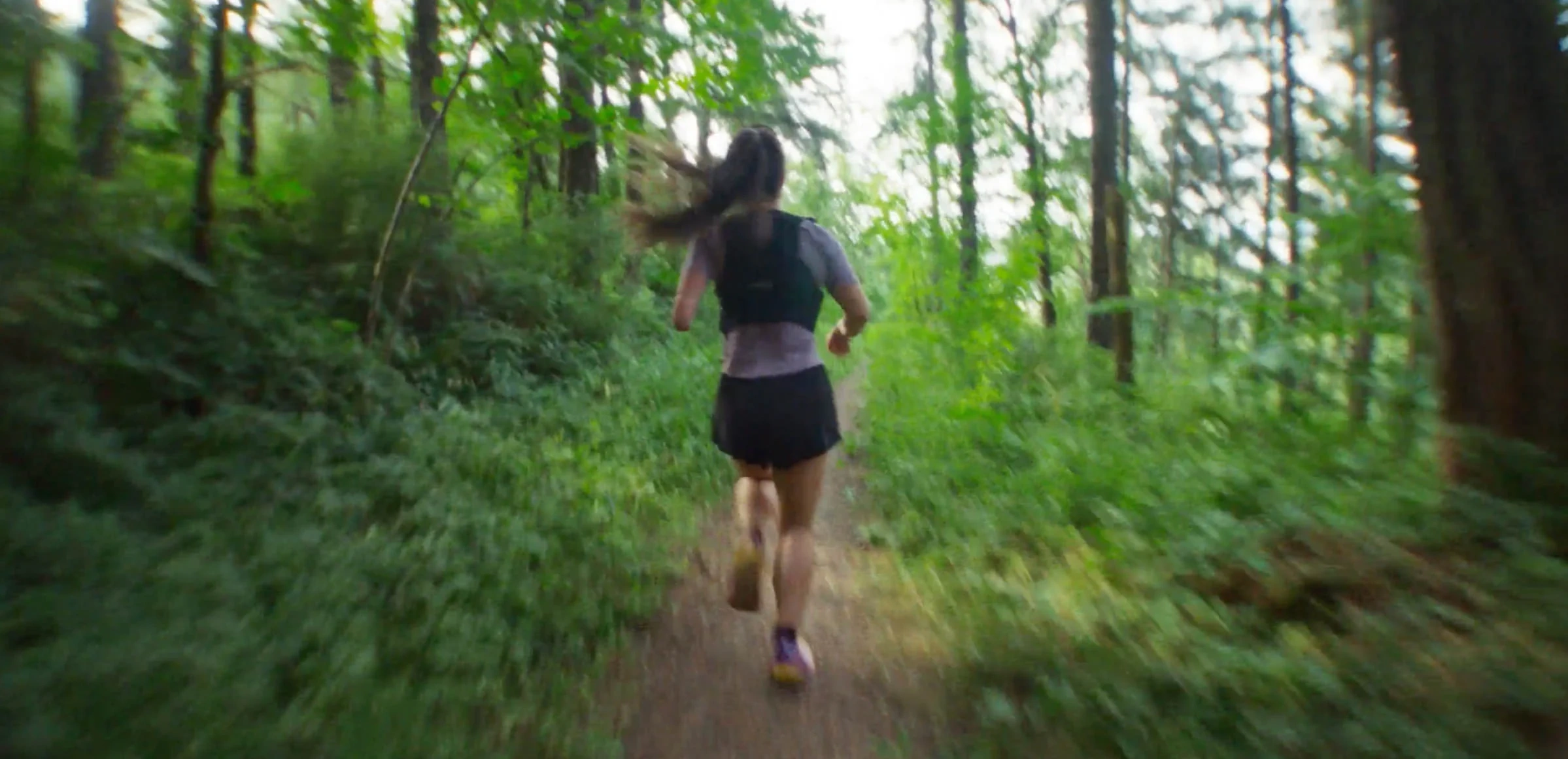 Shot from behind as woman in running vest and long ponytail runs through a dense forest trail.